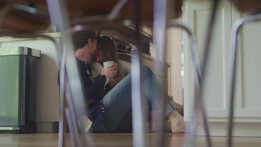 Woman with a striped shirt and man wearing a beanie white sweater and jeans sit on the ground and drink coffee in the kitchen