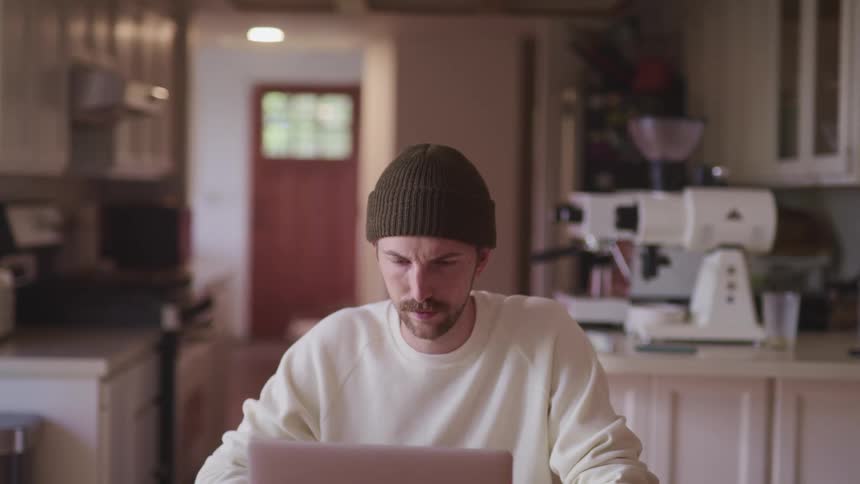 Man with a dark green beanie and a white sweater sits at a table and uses a laptop in the kitchen
