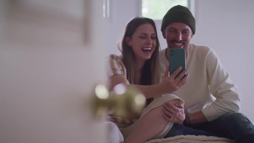 A woman and man are sitting on a bed and taking selfies