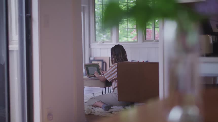 A woman sits on a blanket on the floor working on a laptop on a low table