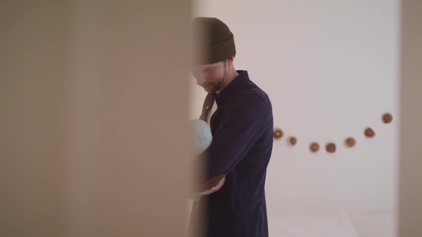 A man can be see cradling a baby while standing next to a lamp through a small opening in the doorway