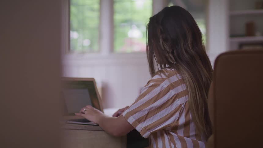 A woman sits at a desk and uses a laptop inside a house during the day