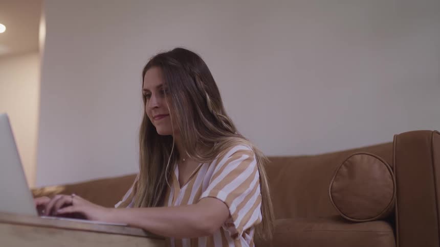 A woman sitting next to a couch in front of a table typing on a laptop