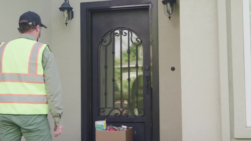 A man delivers groceries on to a step and a woman answers the door and smiles and waves at him