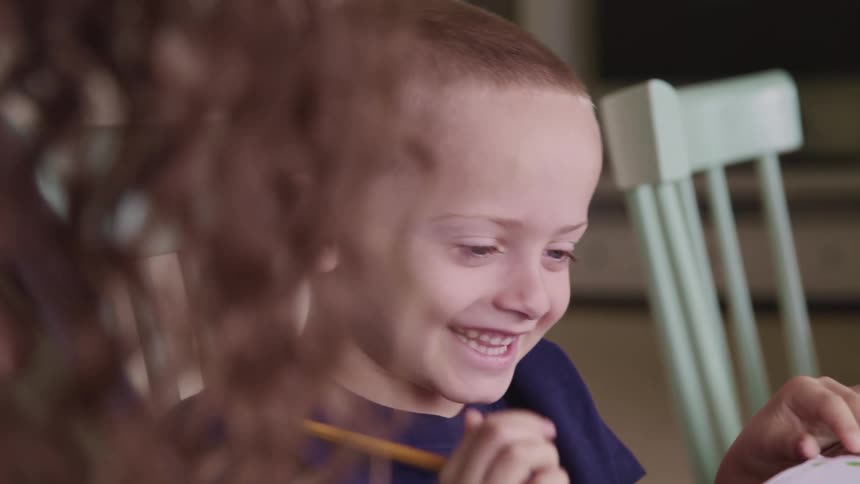 A little boy sits at a table with others and laughs as he draws on a paper