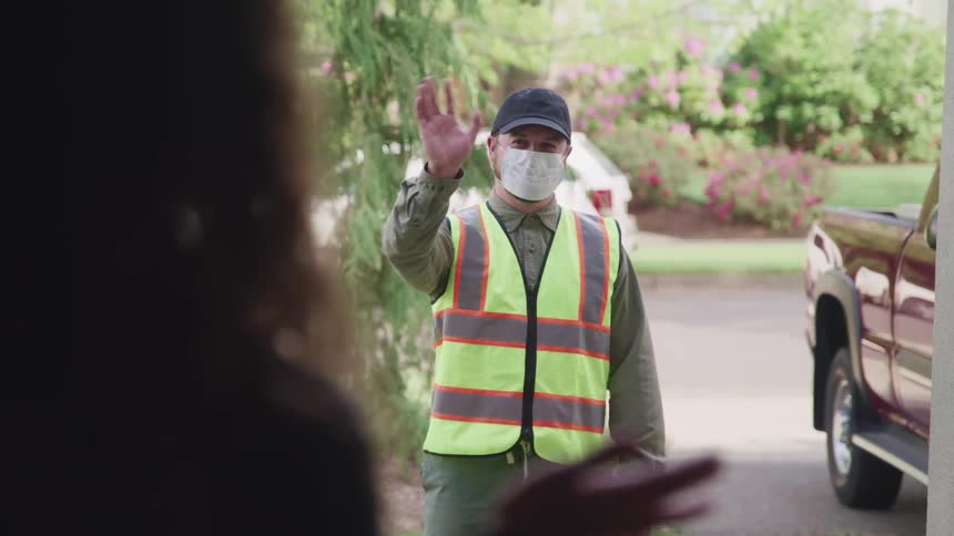 A man stands outside of a home wearing a caution vest and medical mask waving to the person standing inside the home
