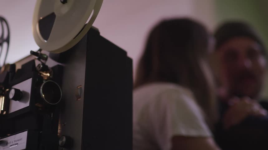 Reels spin on a projector as a man and woman kiss nearby