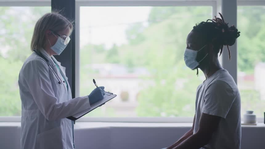 A male patient sits while a medical professional writes notes on a paper on her clipboard