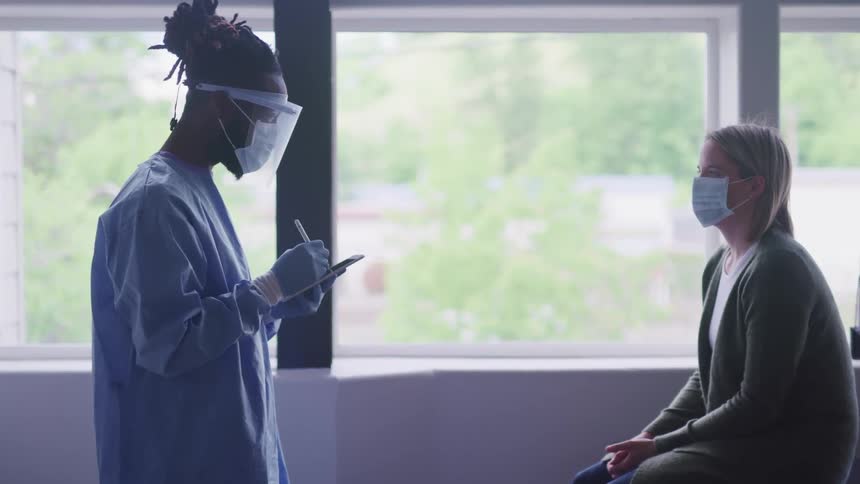 A male nurse in personal protective equipment writes on a notepad while standing across from a female patient in a face mask