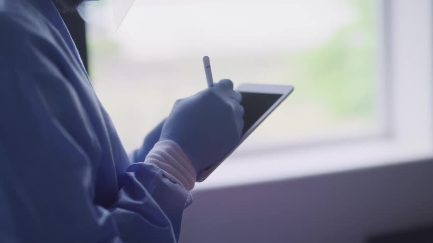 Person wearing blue scrubs and latex gloves writes on a tablet with a stylus
