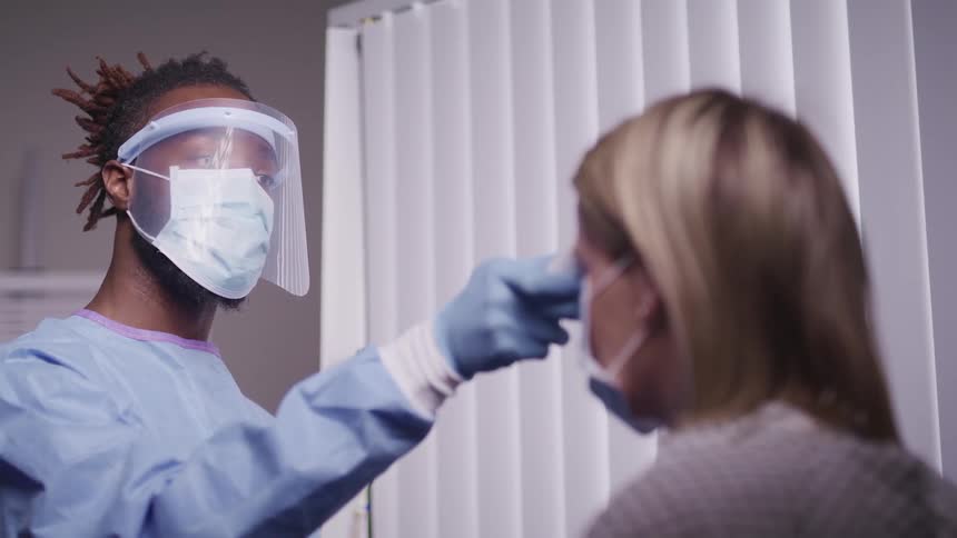Male nurse wearing a face mask and face shield swipes a thermometer across a female patients forehead in an examination room at the hospital