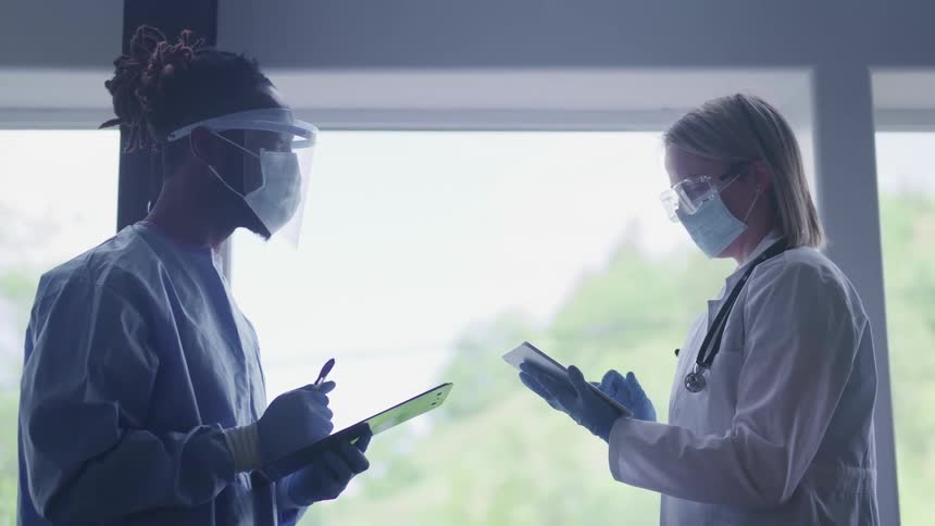 Doctors in lab coats wearing face masks talk to each other taking notes on tablets