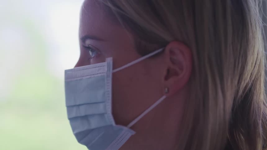 A woman with blonde hair and wearing a disposable face mask slowly raises her gaze