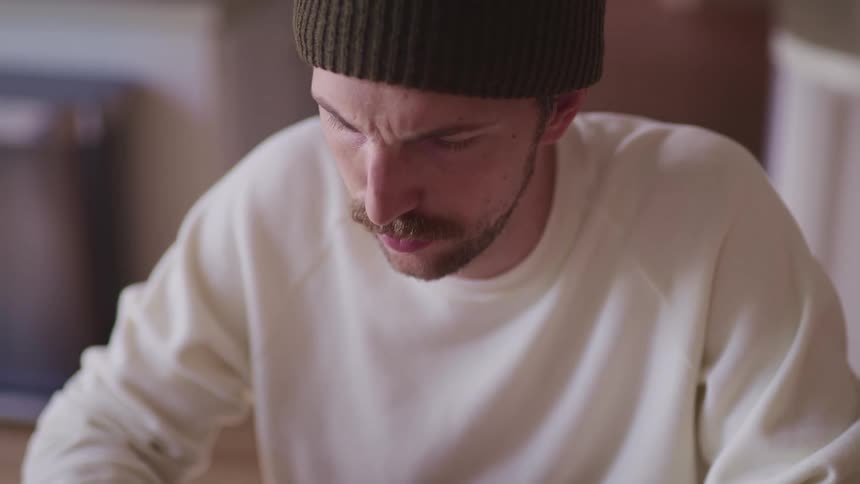 Man with a dark green beanie and a white sweater sits at a table and uses a laptop in the kitchen