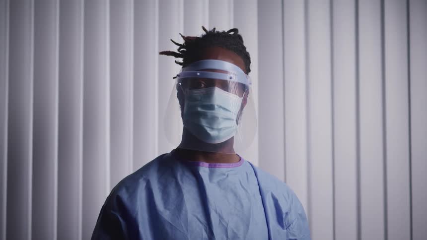 A man with a mask a face shield and hospital gown looking up with window blinds behind him