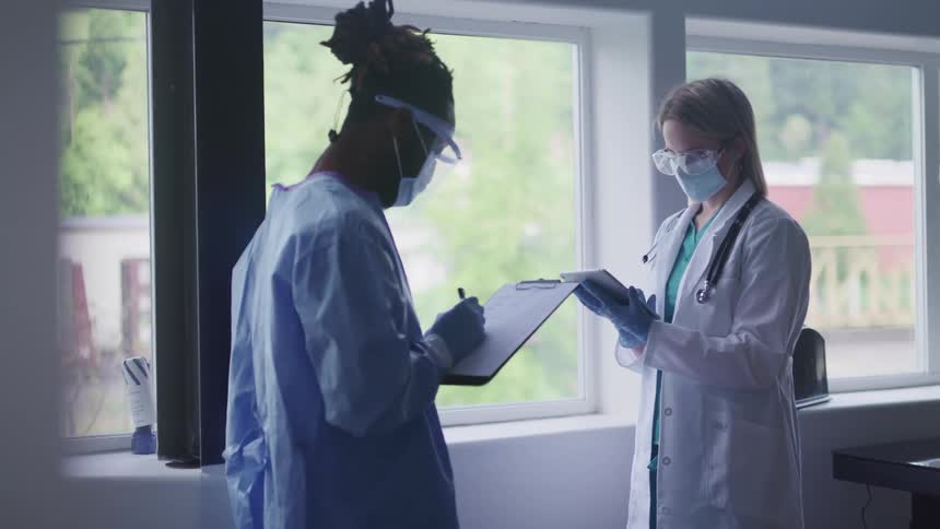 Male nurse wearing ppe in a medical clinic writes on a form on a clipboard as a female doctor stands facing him while typing on a tablet