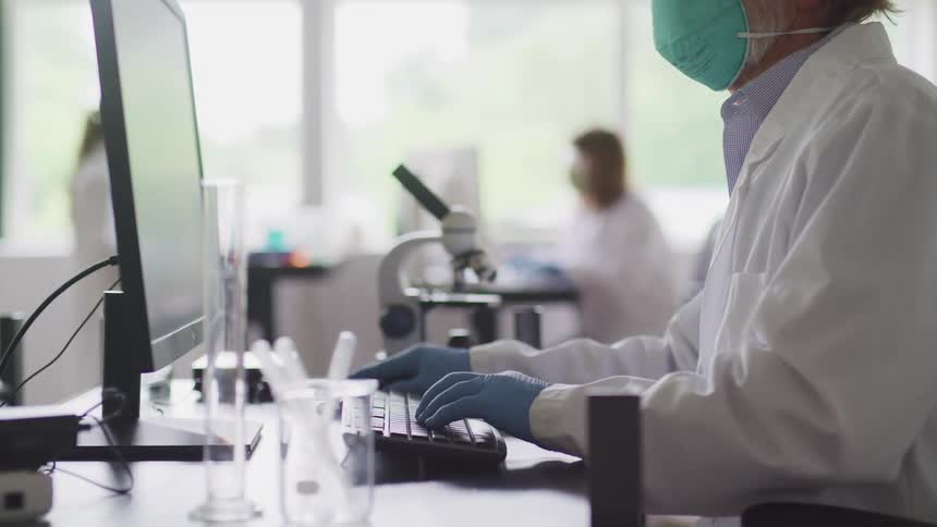 A lab worker types on a computer on a desk covered with test tubes and other equipment