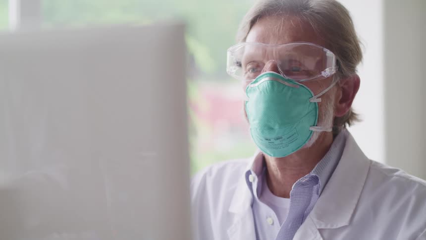 Doctor wearing with a light blue face mask a white lab coat and goggles sits at the desk and uses a desktop