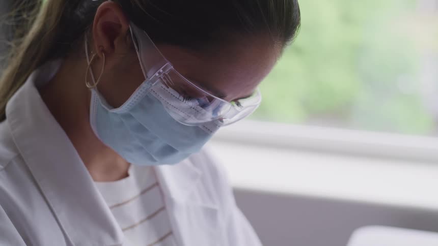 Woman wearing white coat and face mask works near window