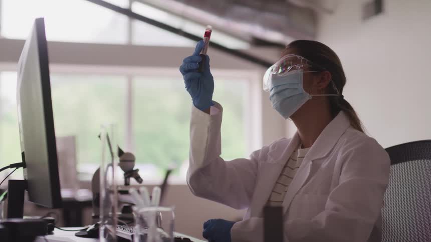 A woman sits at a desk and turns a vile of blood while sitting in front of a computer monitor