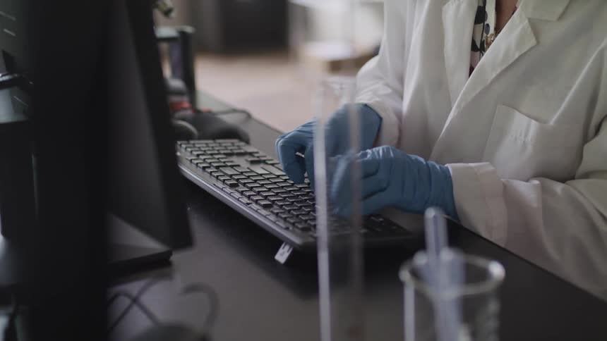 A person with gloves and a lab coat typing on a computer in a lab