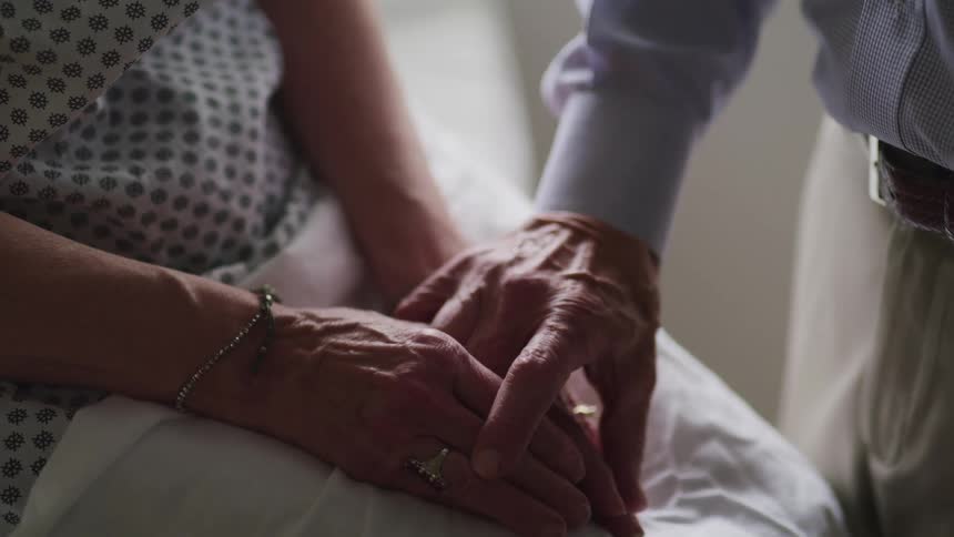 Elderly man reaches for the hand of an elderly woman who is in bed