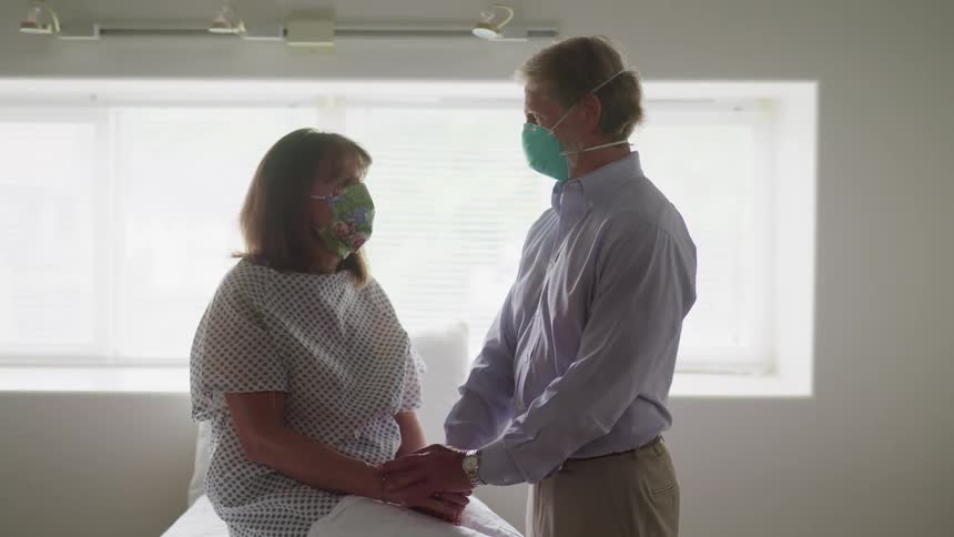 Female patient sitting on examination table in doctors office wearing a patient gown and face mask is being consoled by husband as he reaches for and holds both of her hands