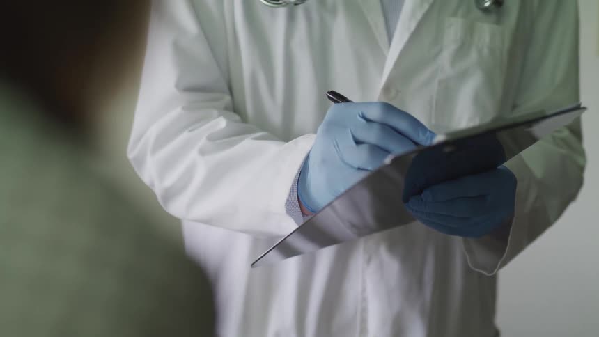 A doctor stands in front of a patient and writes on a chart