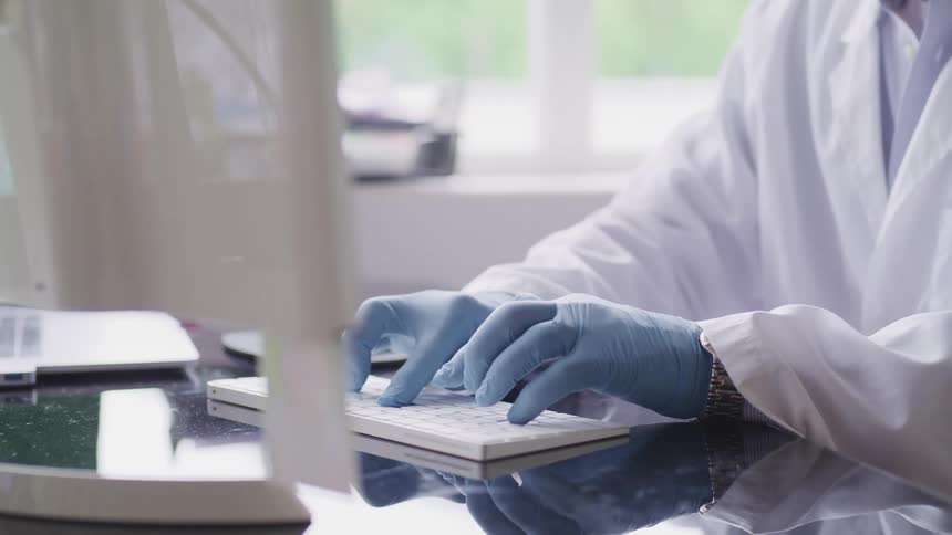 A person with gloves and a lab coat typing on a computer with a window in the background