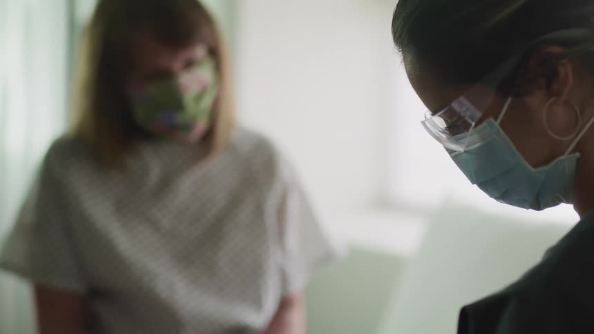 A nurse with a mask and goggles looking down while a patient with a mask sits in the background