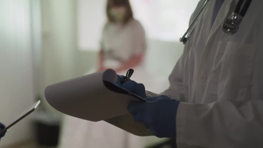 A doctor writes on a clipboard with a pin next to another doctor using a tablet as a patient looks on from her seat across the room