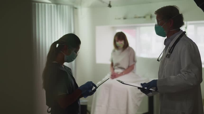 A doctor with a clipboard talking to a nurse with a tablet with a patient in the background