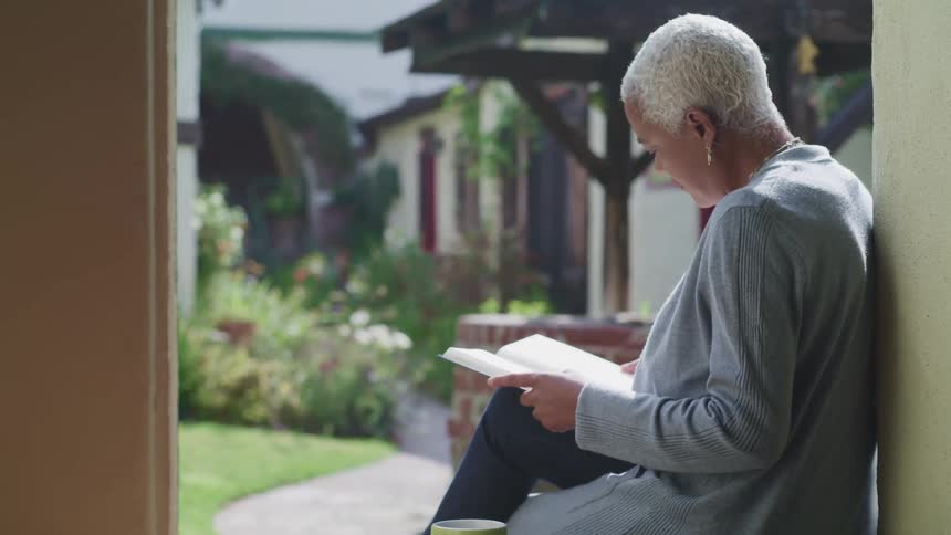 A woman sits outside her house reading a book on a sunny day