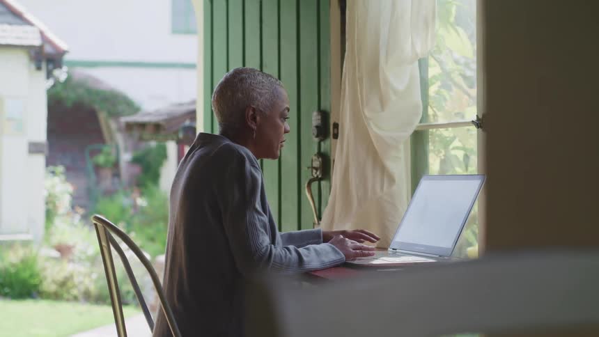 Female sitting a table in front of the window with a view of the garden as she types on the keyboard of the laptop