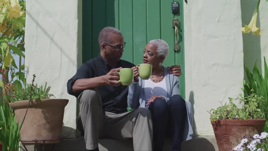 Elderly man and woman sit on the steps holding coffee mugs as they look into each others eyes