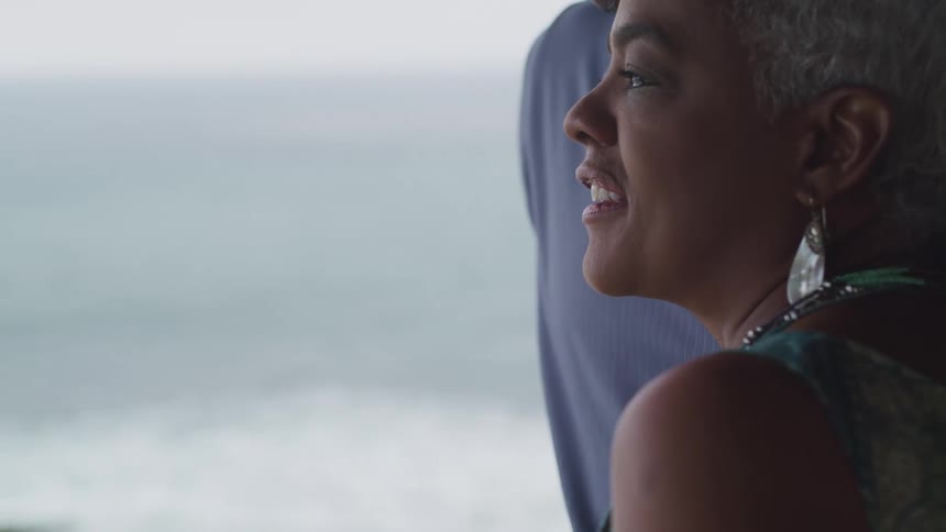 A husband and wife stand near the shore and look at the ocean