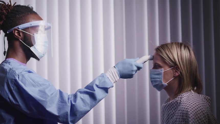 A doctor with a mask and gloves using a thermometer on a woman patient head
