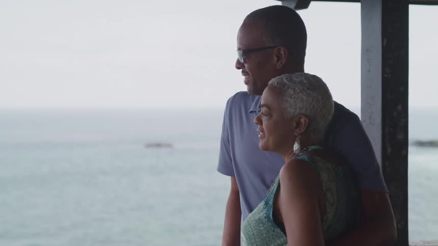 Couple stand on a porch arminarm and look out onto the ocean view while smile