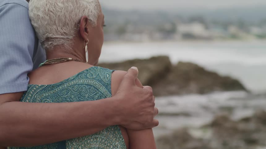 A man embraces an elderly woman as the two stand on the coast and gaze out at the ocean