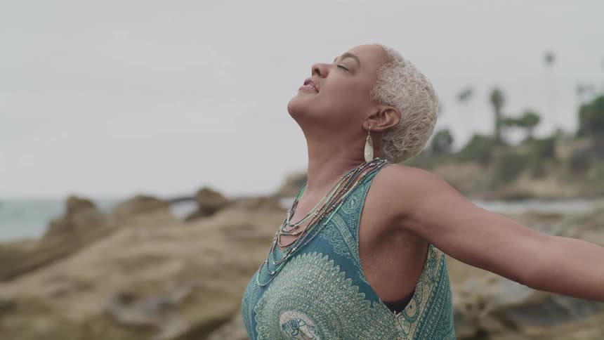 Woman standing arms out with eyes closed facing the sky on a cliff overlooking the beach