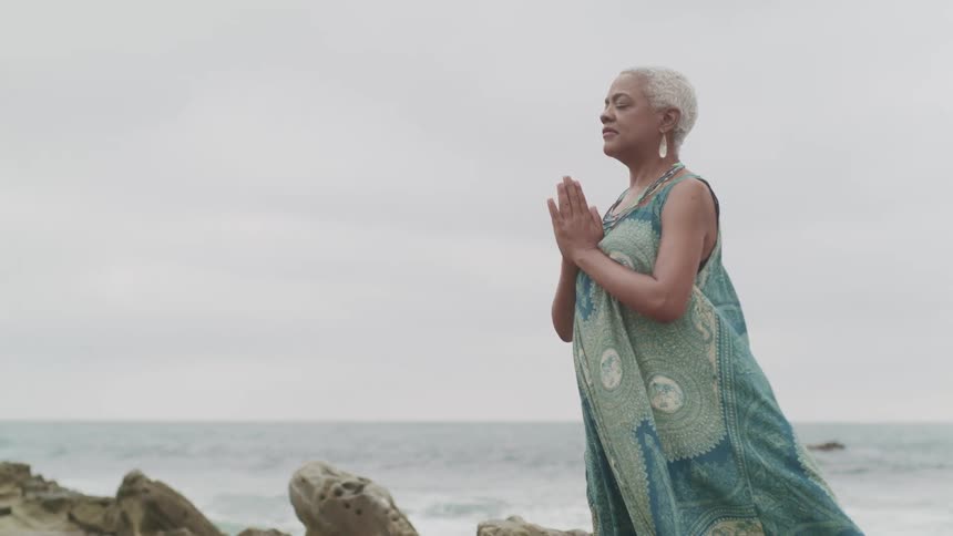 A woman meditates on a rock formation next to the ocean on a cloudy day
