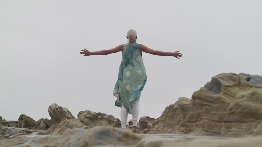 A woman stands on a rocky surface with her arms outspread