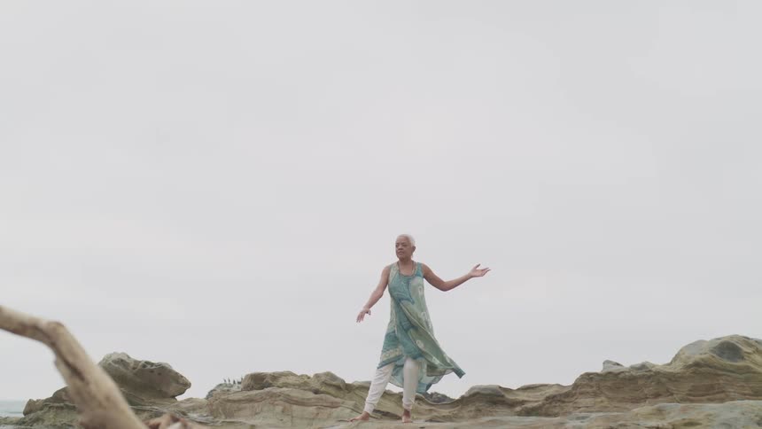 A woman stands on a cliff overlooking the ocean and does tai chi