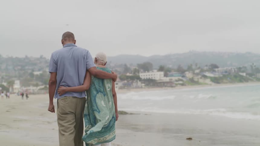 Middle aged black man wearing a blue polo shirt and brown pants and a woman with a grey short hair and green dress walk on the beach by the sea on a windy and cloudy day