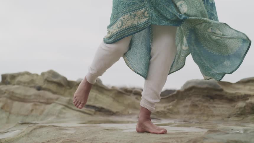 Woman moves through dance poses standing barefoot on stone on windy day