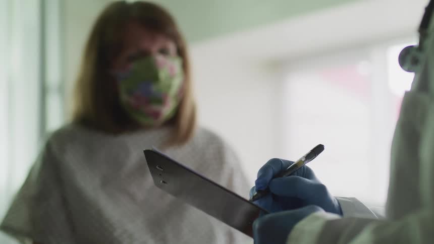 A doctor writes on a clipboard as a patient in a hospital gown and mask watches nearby