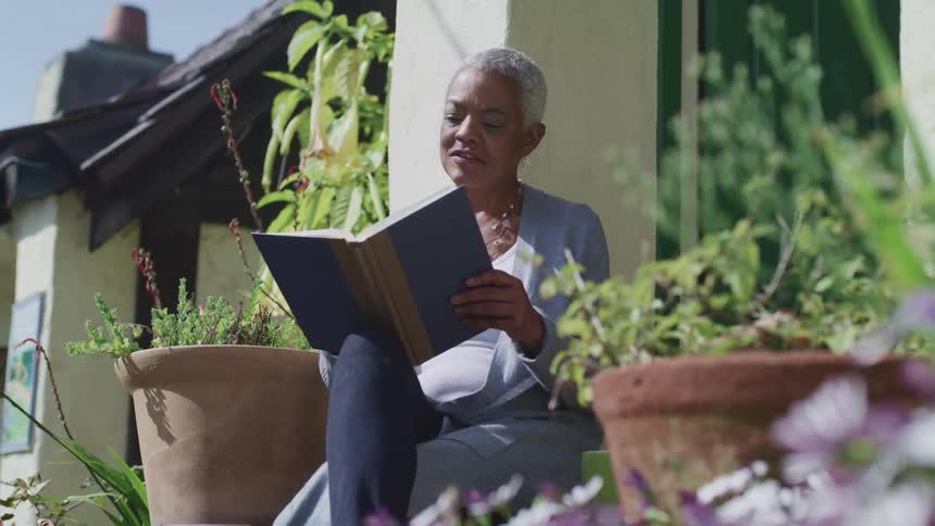 A woman reads a book while sitting surrounded by potted plants outside of a house