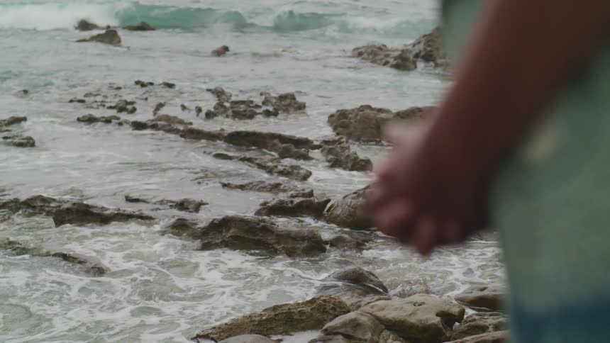 A person stands on a rocky beach with their hands clasped