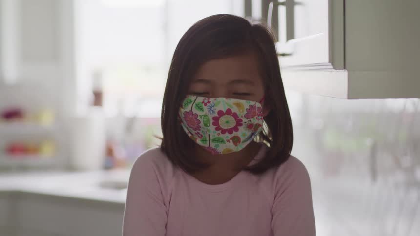 Asian child with straight hair and a face mask looks at the camera in the kitchen