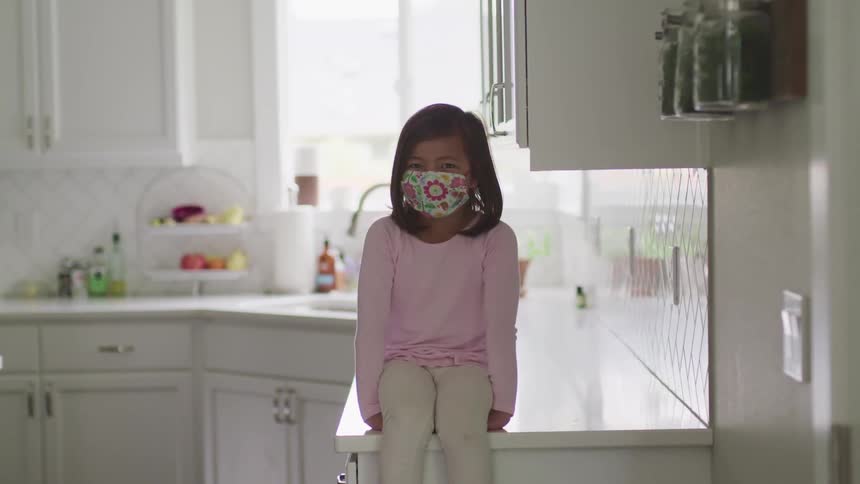 Girl wearing face mask sits on counter in white kitchen smiling slightly
